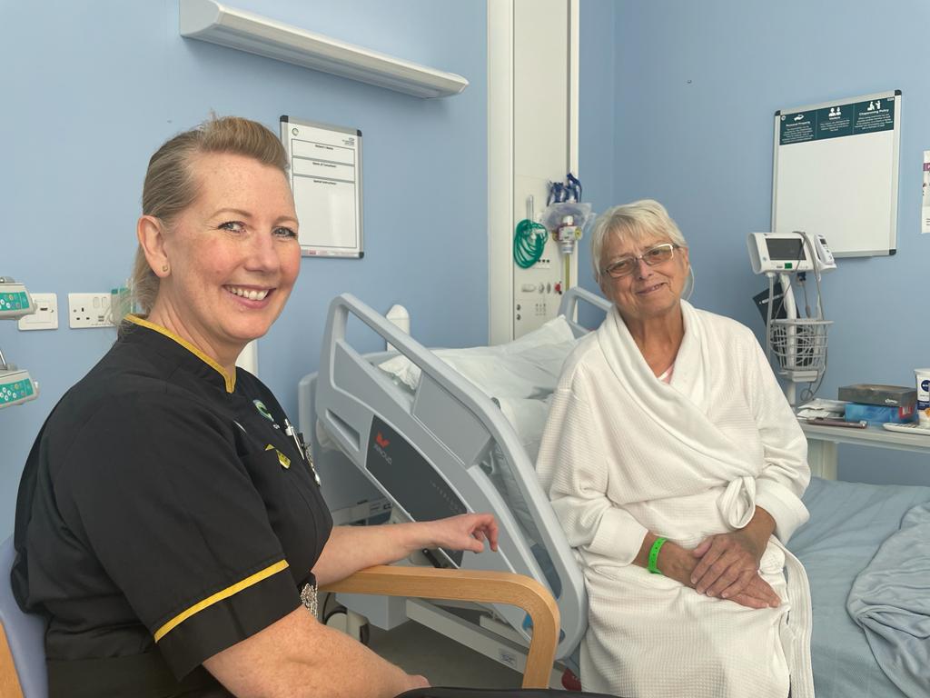 Picture of Julie Gray, who is wearing a dark navy nurses uniform, sitting on a ward chair with a hospital patient who is on her hospital bed and is smiling