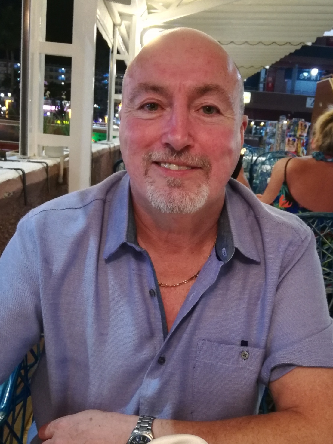 Picture of Graeme Brooks sitting in a restaurant. He is wearing a light blue short sleeved shirt and has a short grey beard