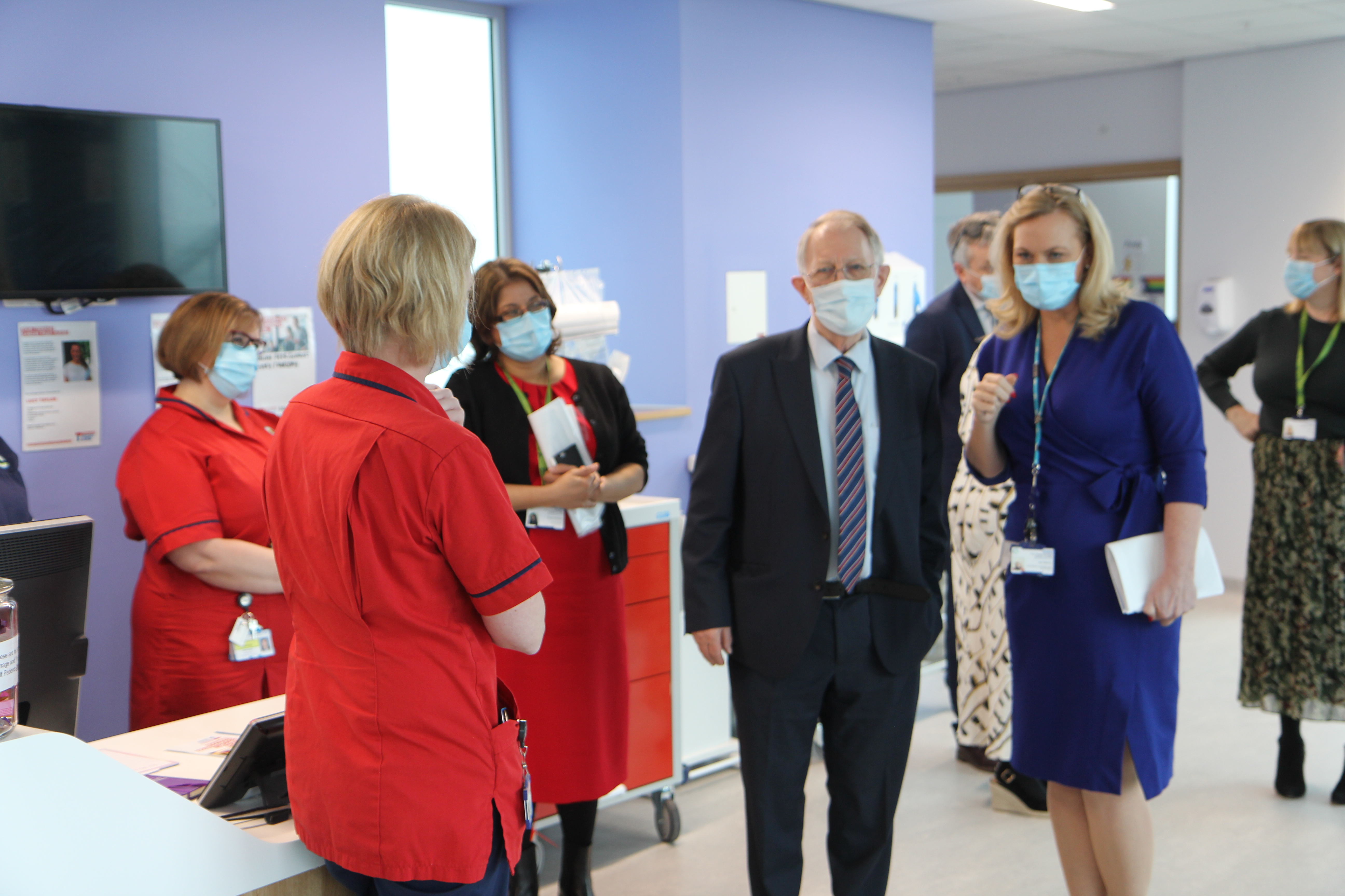 Professor Sir Mike Richards is pictured on the Teenage & Young Adult Unit with hospital staff including nurses in red uniforms. They are chatting and look relaxed and happy. 