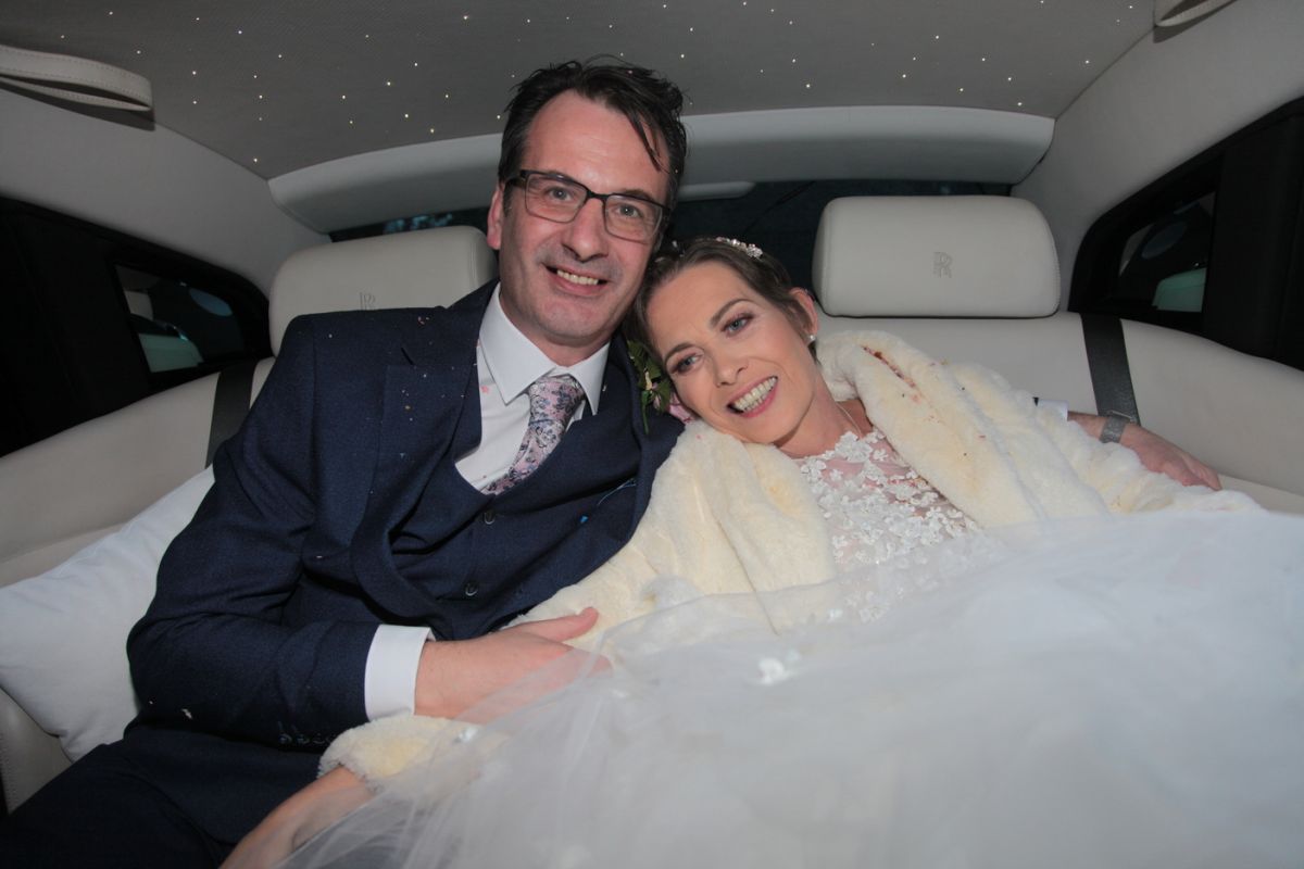 A bridegroom and bride in the back of a beautiful car after their wedding. The man has glasses and is wearing a black dress suit. The woman has a beautiful white wedding dress, furry stole and veil. They are leaning in to each other and look really happy.