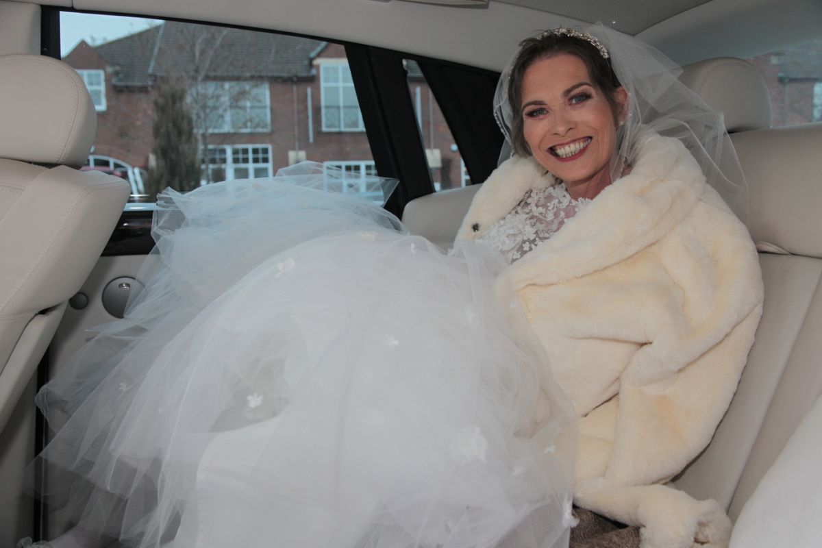 A woman sitting in the back of a beautiful car. She is wearing a gorgeous white wedding dress and veil and has a furry stole around her shoulders. She is smiling and looks really happy. 